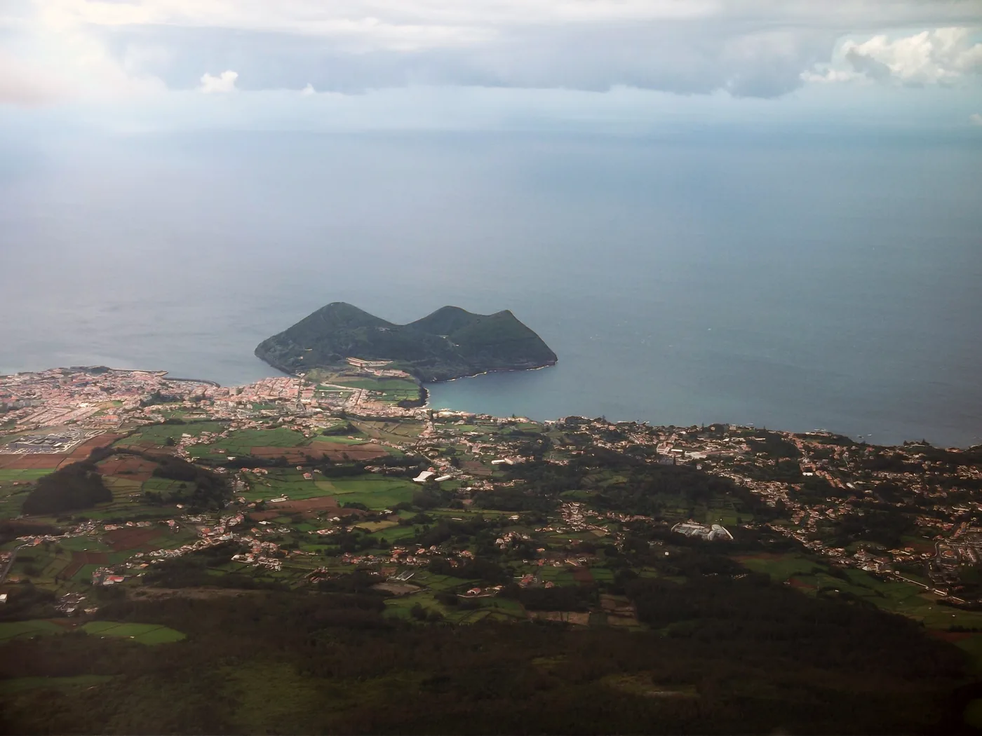 An aerial view of a town spread along a bay, with a dark volcanic peninsula sheltering its harbour and farmland behind.