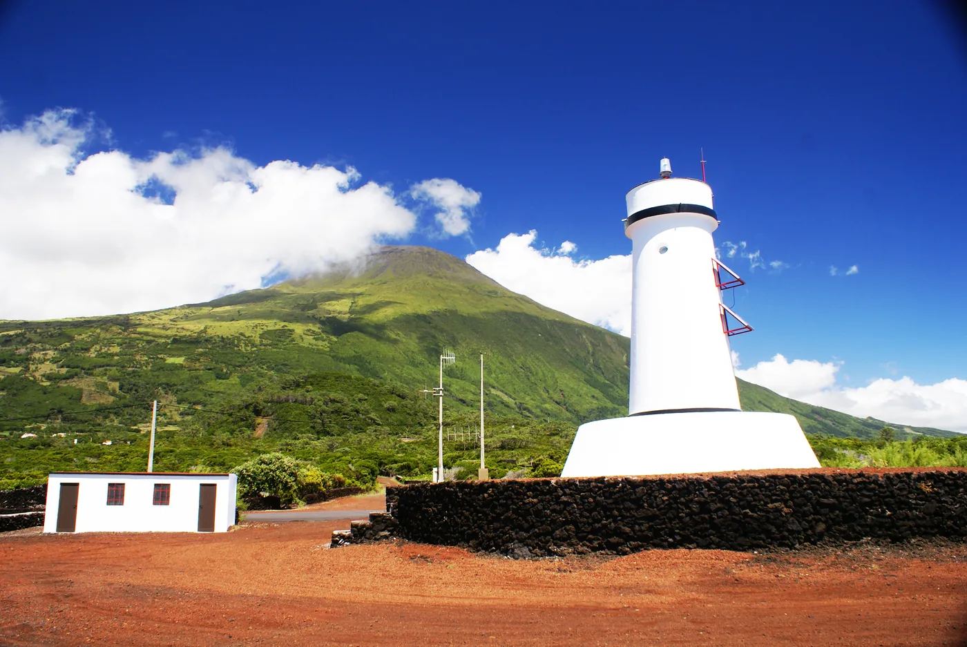 A white lighthouse behind a low lava-stone wall on red volcanic ground, with the green cone of Pico mountain rising into cloud behind it.