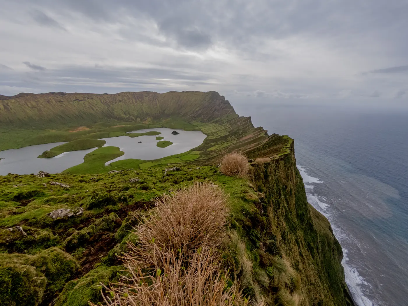 A broad grassy crater with small lakes on its floor, seen from the rim, with the island’s cliffs and the sea beyond.