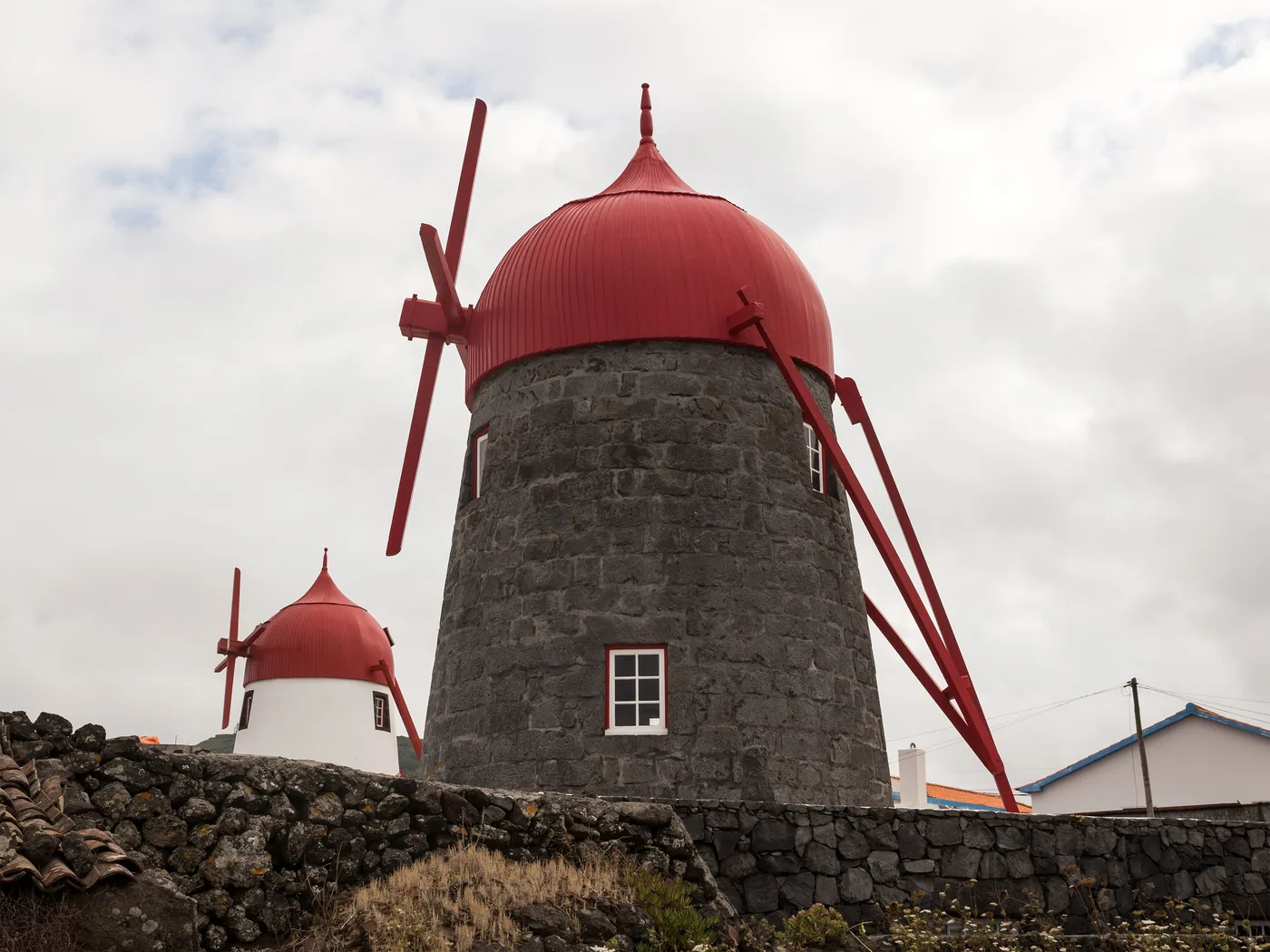 Two stone windmills with red domed caps and red sails, beside a lava-stone wall and white houses under a cloudy sky.