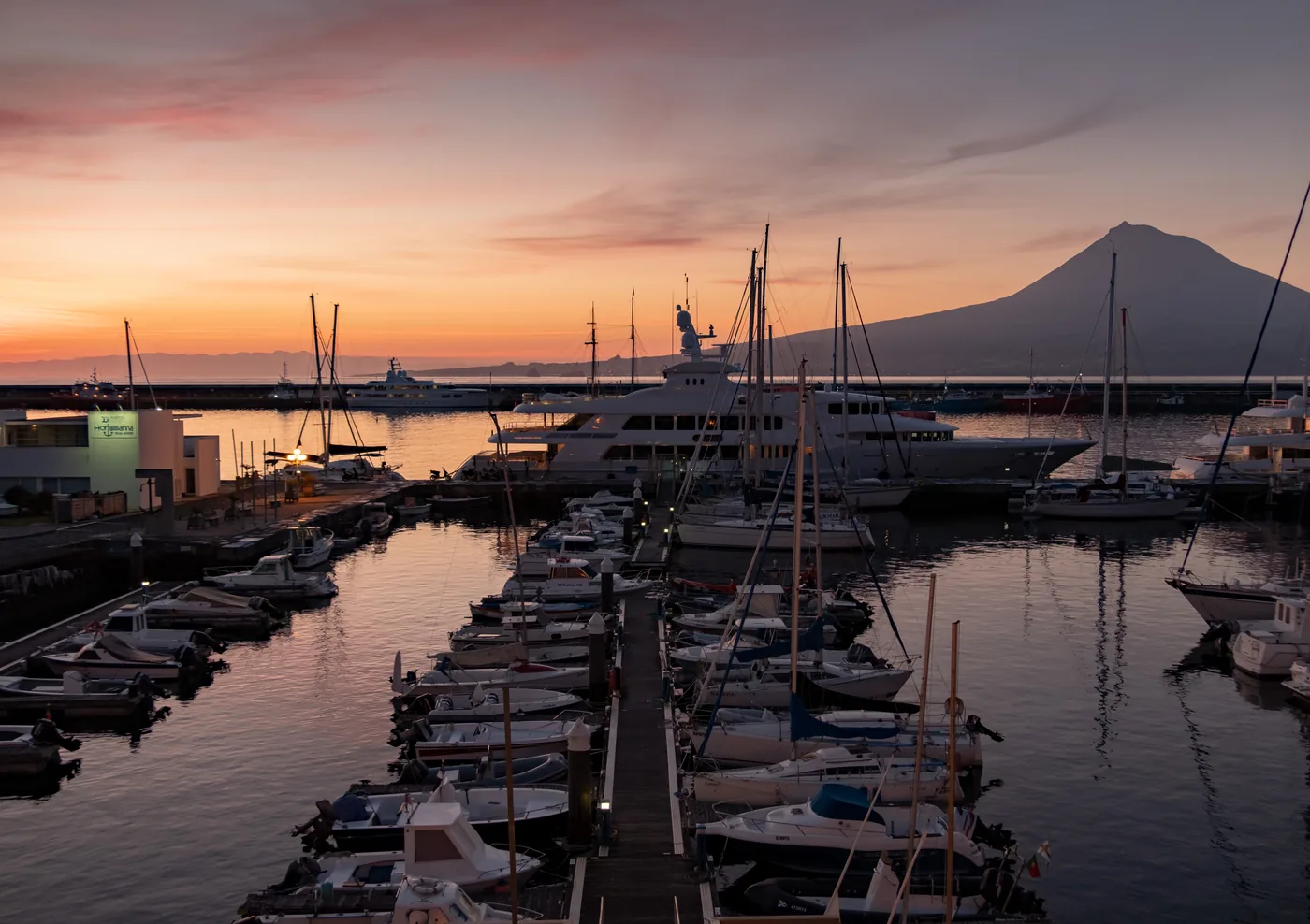 Yachts moored in a marina at sunrise, with painted murals on the quay and a town rising on the hillside behind.