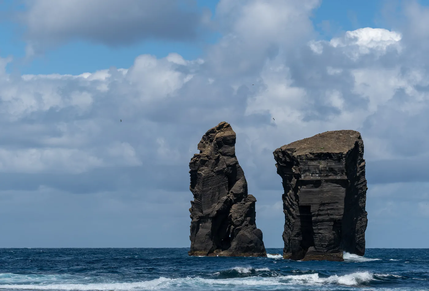 Two tall black sea stacks standing in rough blue water close to a rocky shore under a clear sky.