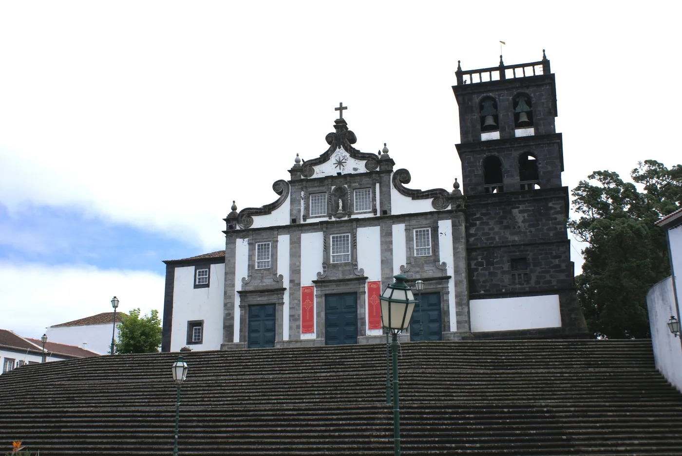 A white baroque church front with dark basalt trim and a square bell tower, at the top of a wide flight of stone steps.