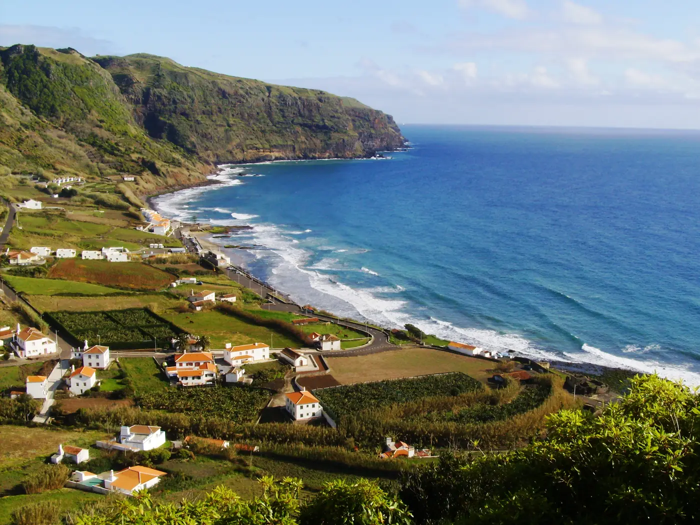 A sandy bay below green cliffs, with white houses and vineyards on the slope above and surf breaking on the beach.