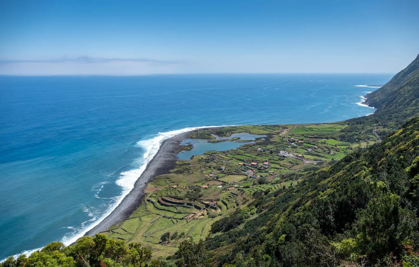 A flat coastal shelf with a lagoon and small fields at the foot of steep green cliffs, seen from above with the open sea beyond.