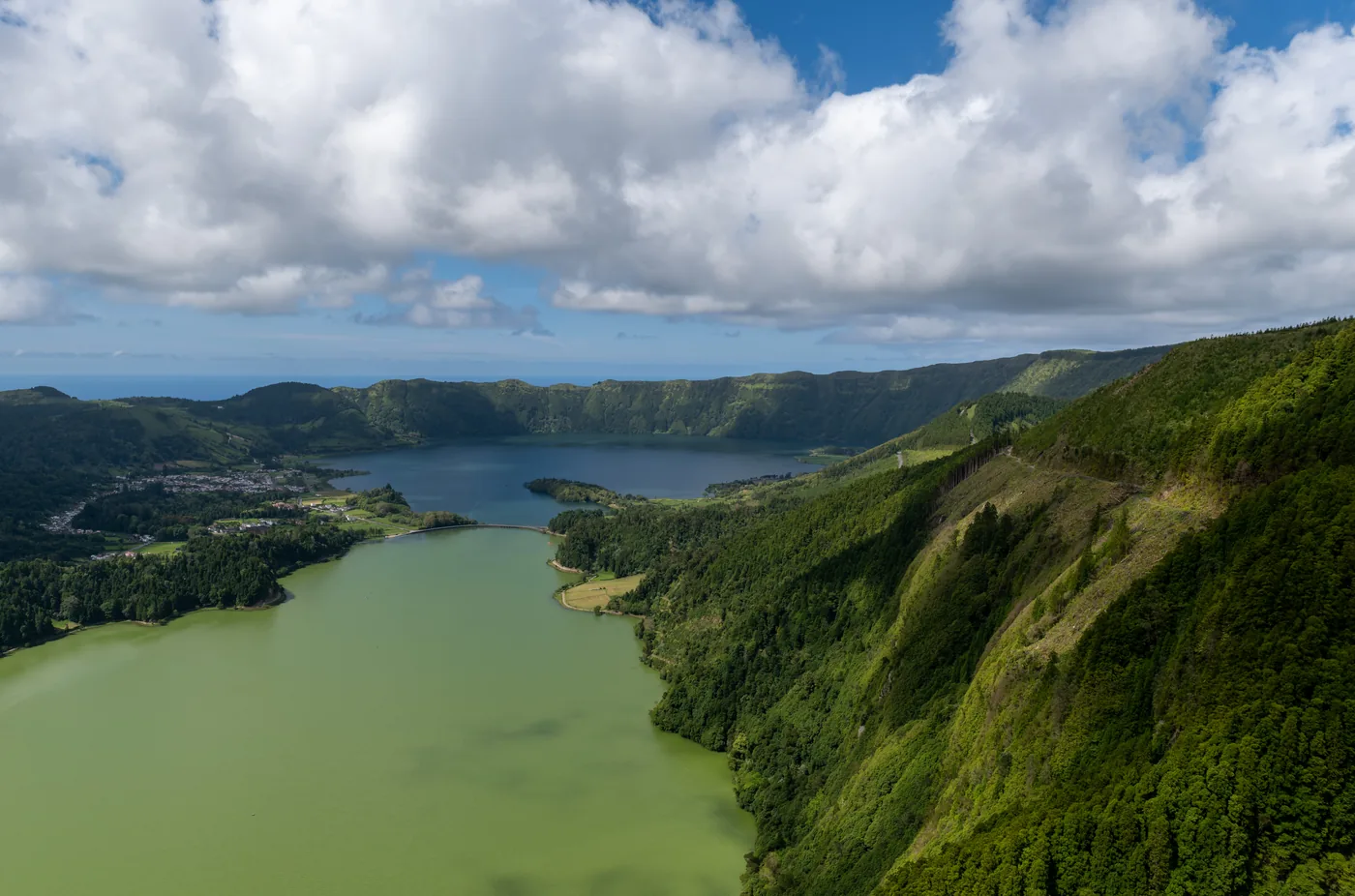 Two crater lakes, one green and one blue, seen from the caldera rim with a village between them and the far crater wall under cloud.