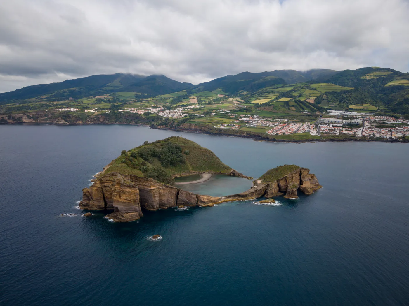 A round volcanic islet with a lagoon in its centre, seen from the air off a coastal town with green hills behind.